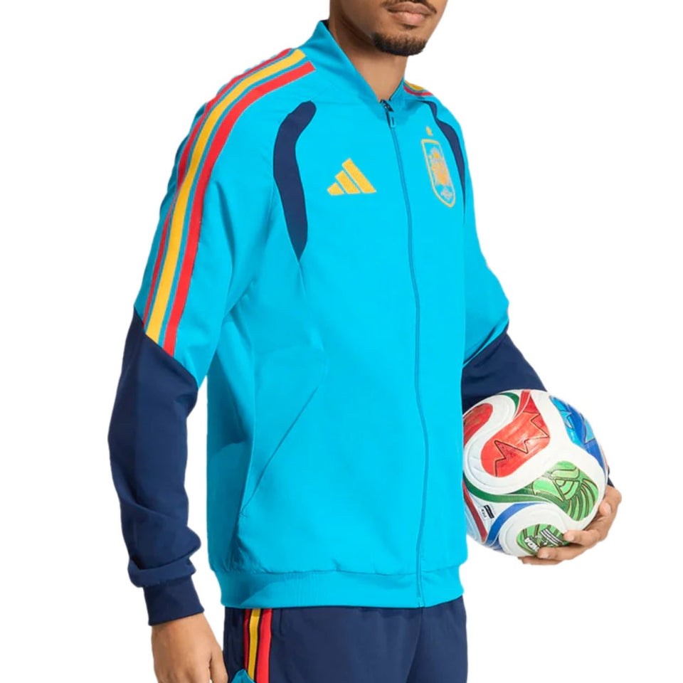 Person wearing a blue Adidas x Spain tracksuit with colorful stripes, holding a soccer ball on a white background