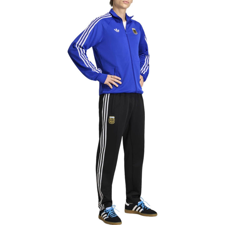 Blue Adidas tracksuit jacket with white stripes and black pants featuring an emblem on the leg.